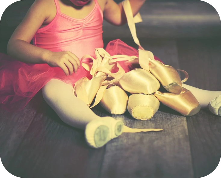 A little girl in pink dress sitting on the floor with ballet shoes.